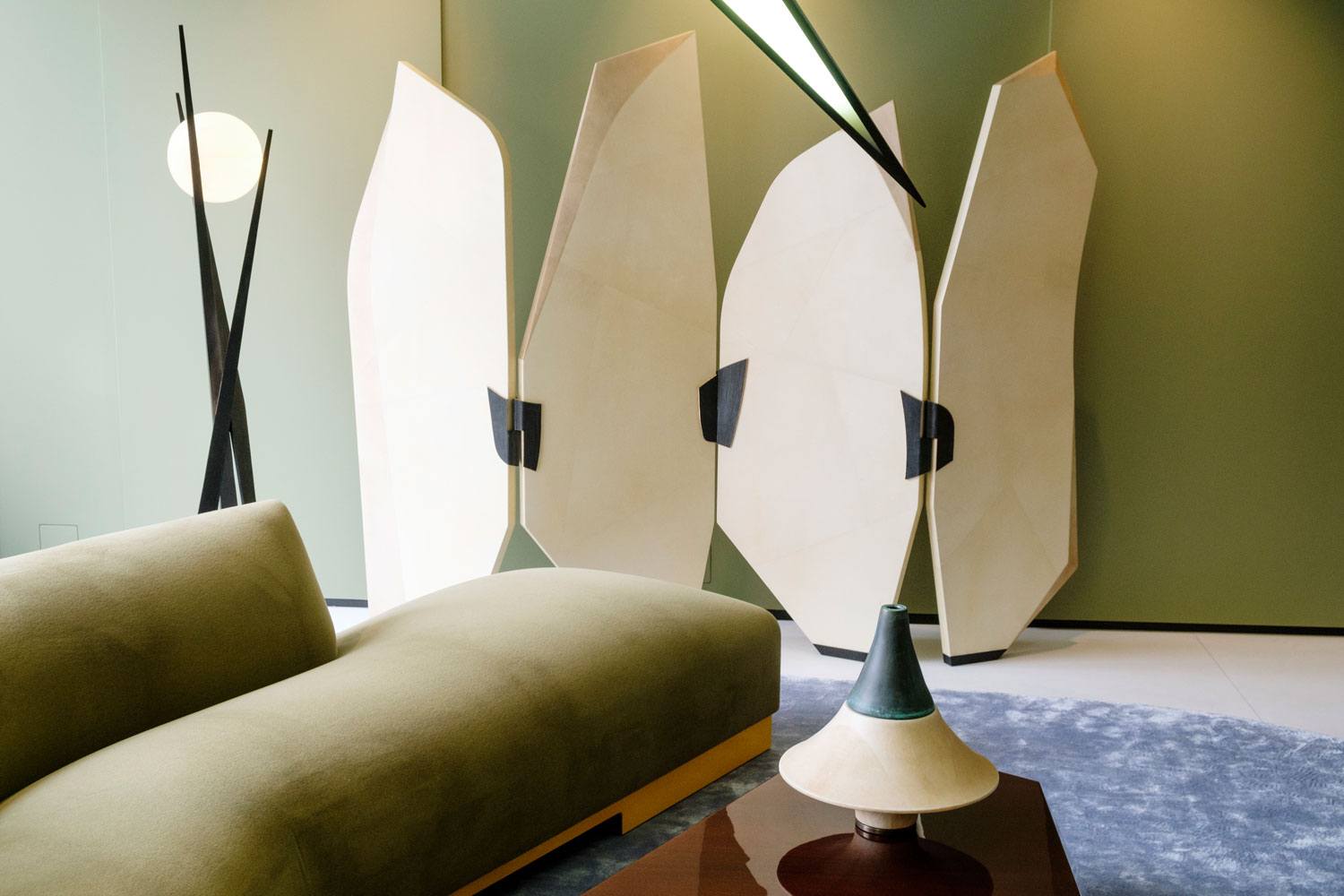 Modern living room with minimalist design, featuring a green couch, abstract room divider, geometric lamp, and neutral tones.