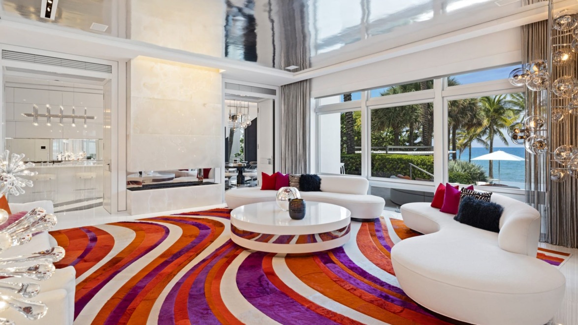 Modern living room with retro colorful rug, white curved sofas, large windows, and view of palm trees and ocean.