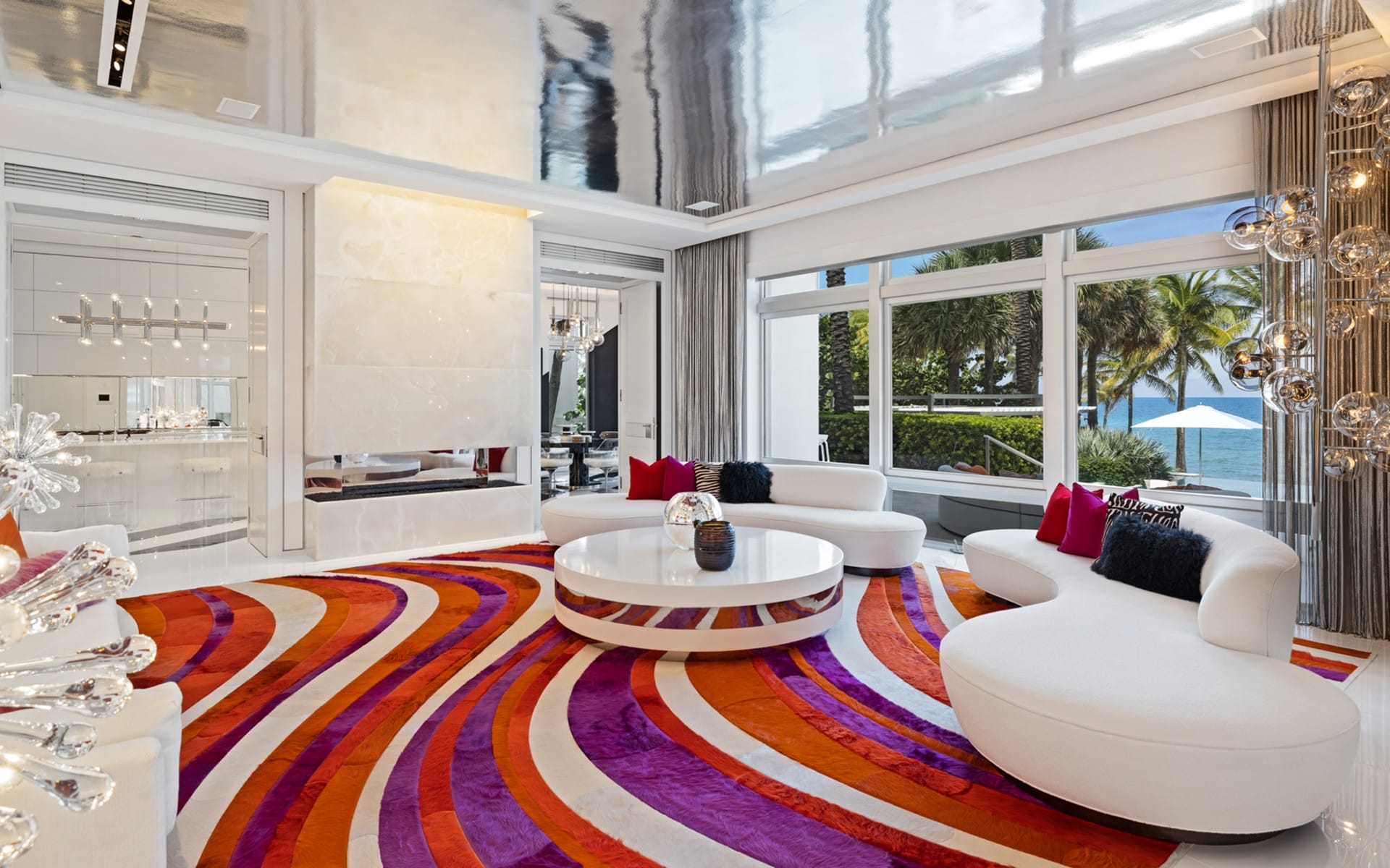 Modern living room with retro colorful rug, white curved sofas, large windows, and view of palm trees and ocean.