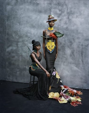 Two models in vibrant, artistically designed outfits and hats, posing against a textured gray background.