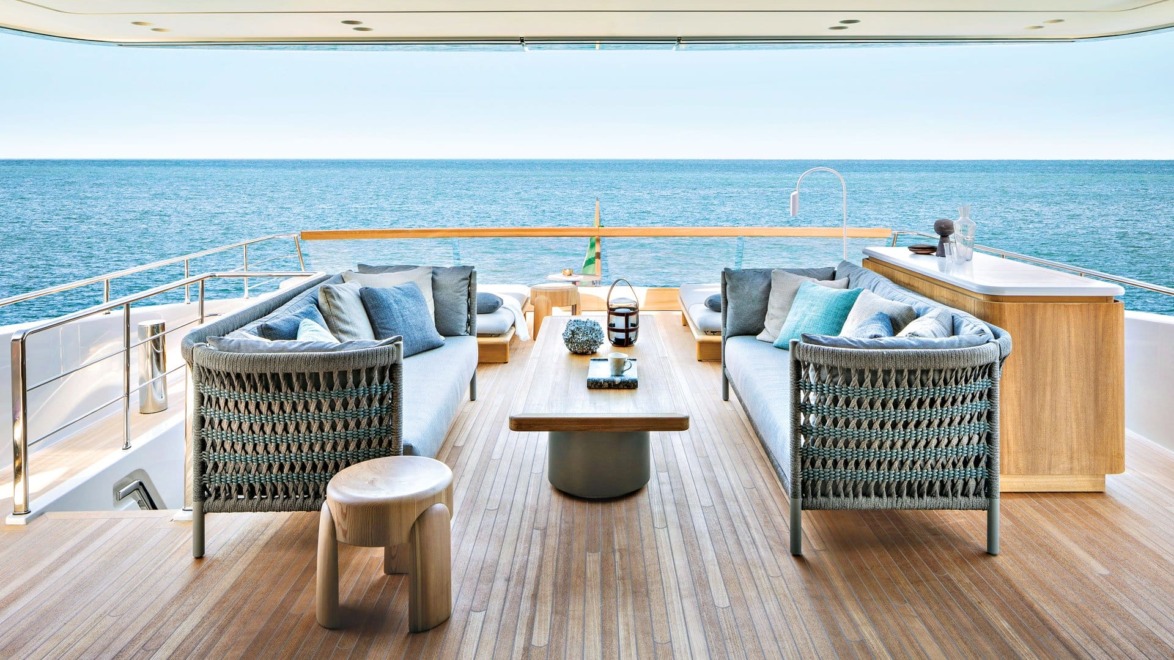 Luxury yacht deck with elegant seating, sea view, and wooden furnishings, featuring cushions and a table, under a clear sky.