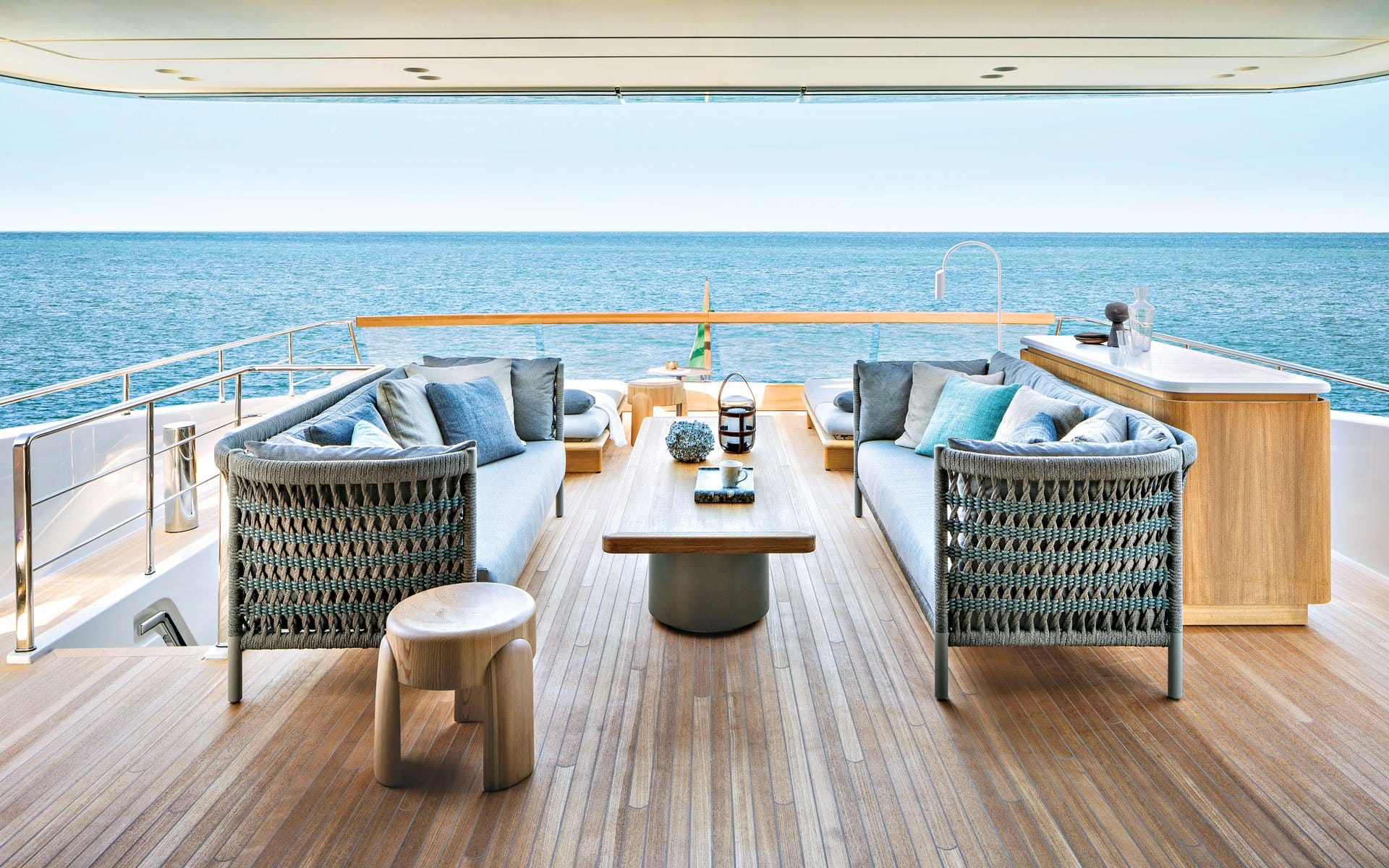 Luxury yacht deck with elegant seating, sea view, and wooden furnishings, featuring cushions and a table, under a clear sky.