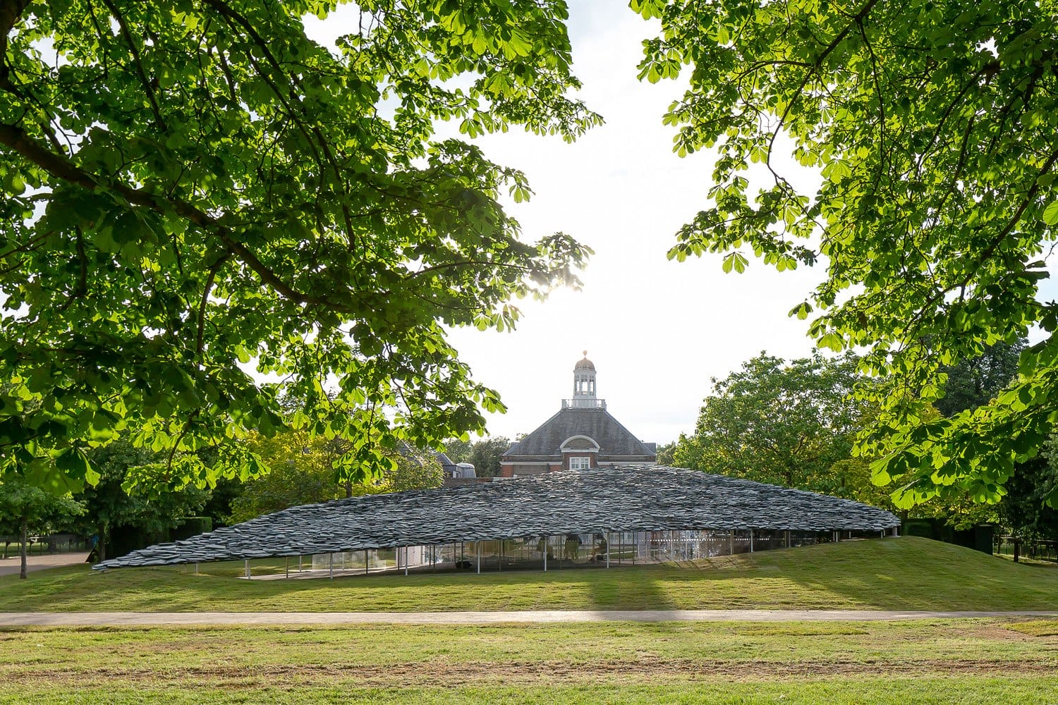 Modern architectural pavilion with a wavy roof in a park, surrounded by lush green trees and a historic brick building in the background.