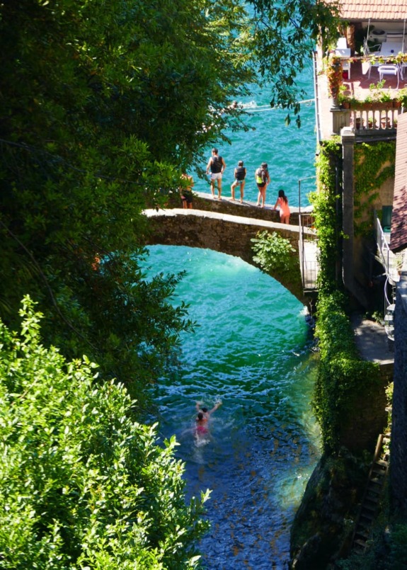 People standing on a stone bridge over a scenic turquoise river with surrounding greenery and buildings in the background.