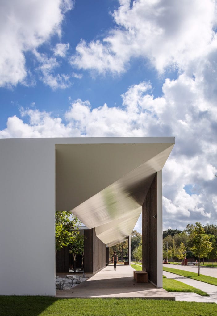 Modern architectural structure with angular roof, surrounded by greenery and cloudy sky, person walking through shaded path.