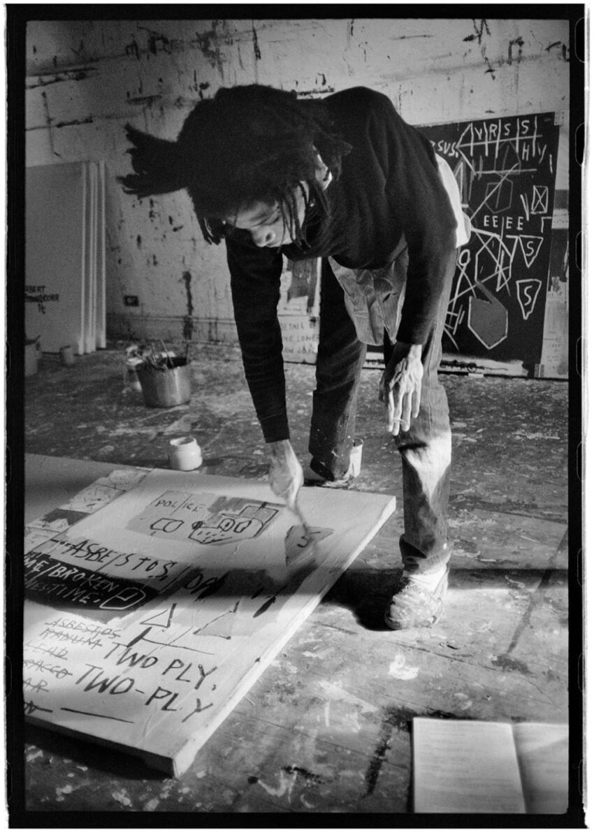 Artist working in a studio, leaning over a canvas on the floor with paint cans in the background.