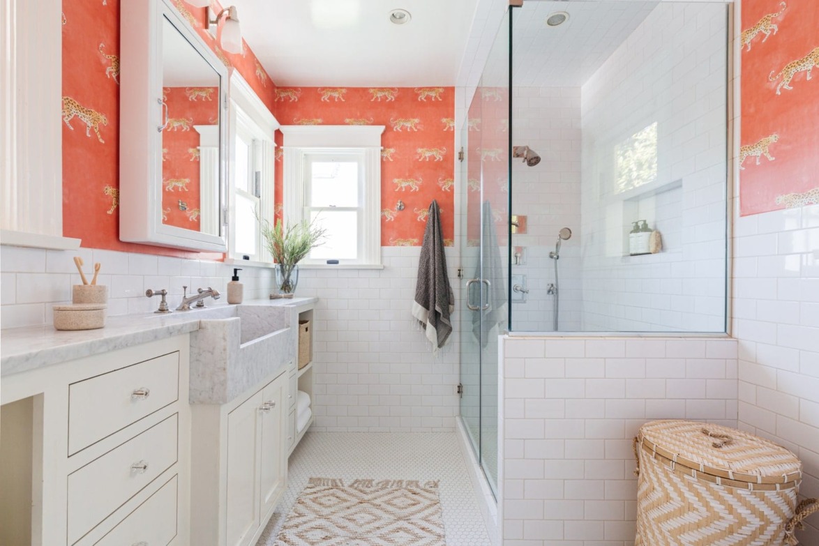 Bright bathroom with glass shower, orange wallpaper with tiger print, white cabinets, and wicker laundry basket.