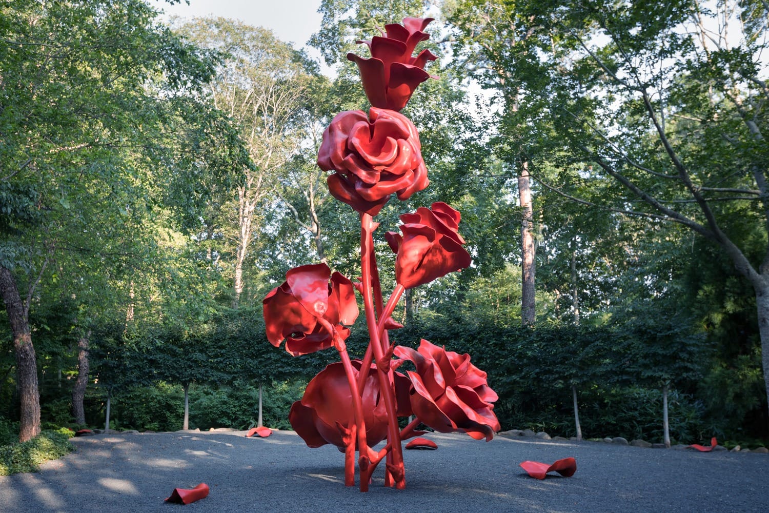 Large red rose sculpture in a serene garden setting, surrounded by greenery and trees, with fallen petals on the ground.