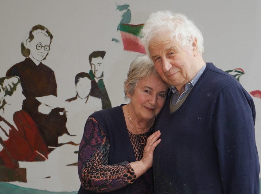 elderly couple embracing in front of an unfinished painting with partially completed figures in the background
