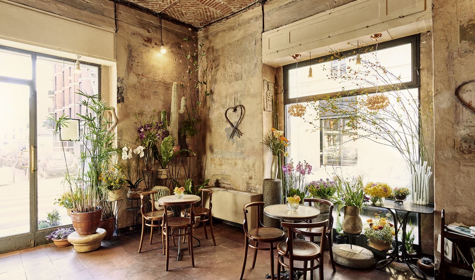 Cozy café interior with rustic decor, wooden chairs, marble tables, and vibrant plants near large windows.