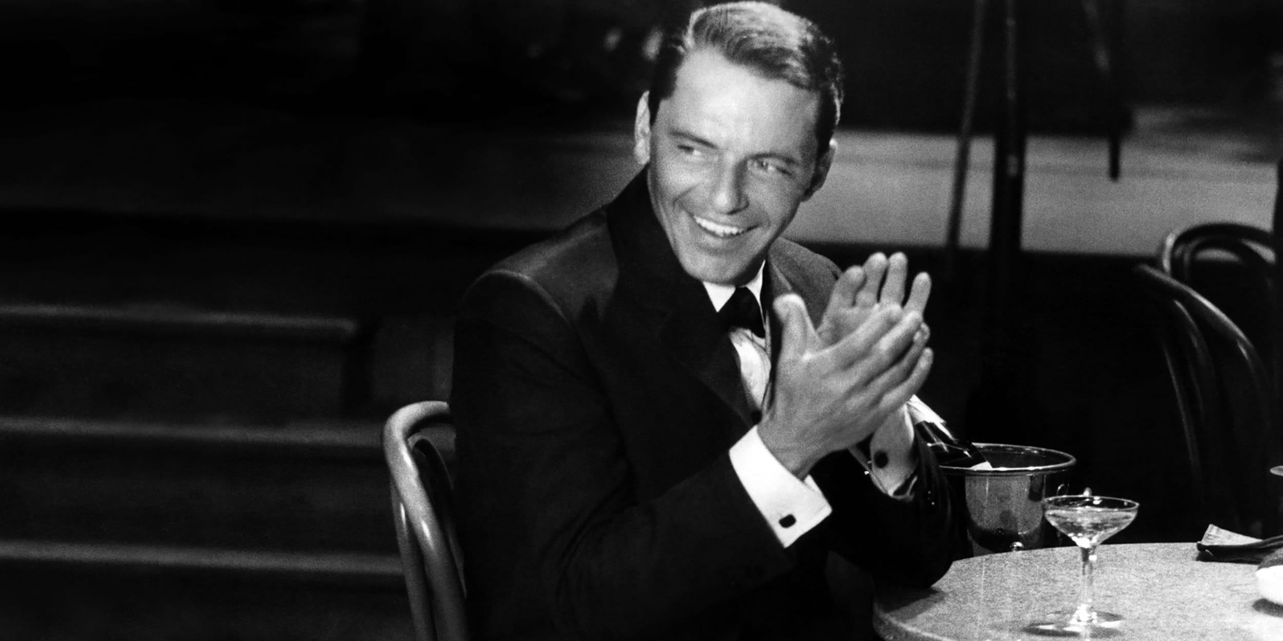 Man in a suit clapping while seated at a table with a champagne glass and bucket.