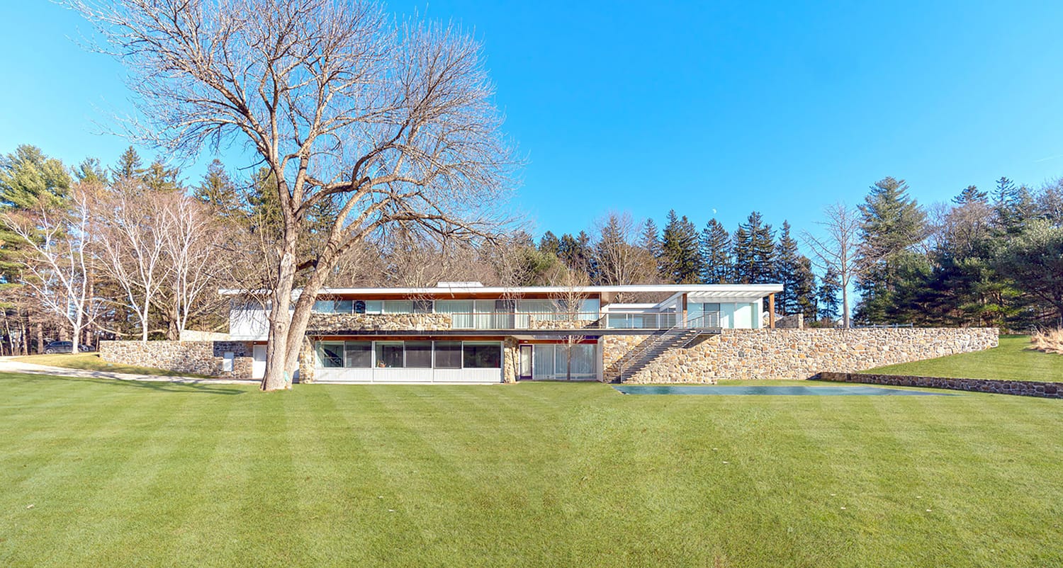 Modern house with glass facade surrounded by trees and a large lawn on a clear day.