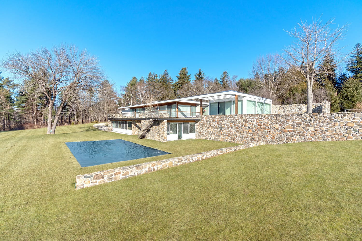 Modern glass and stone house with large yard, surrounded by trees, featuring a reflecting pool on a sunny day.