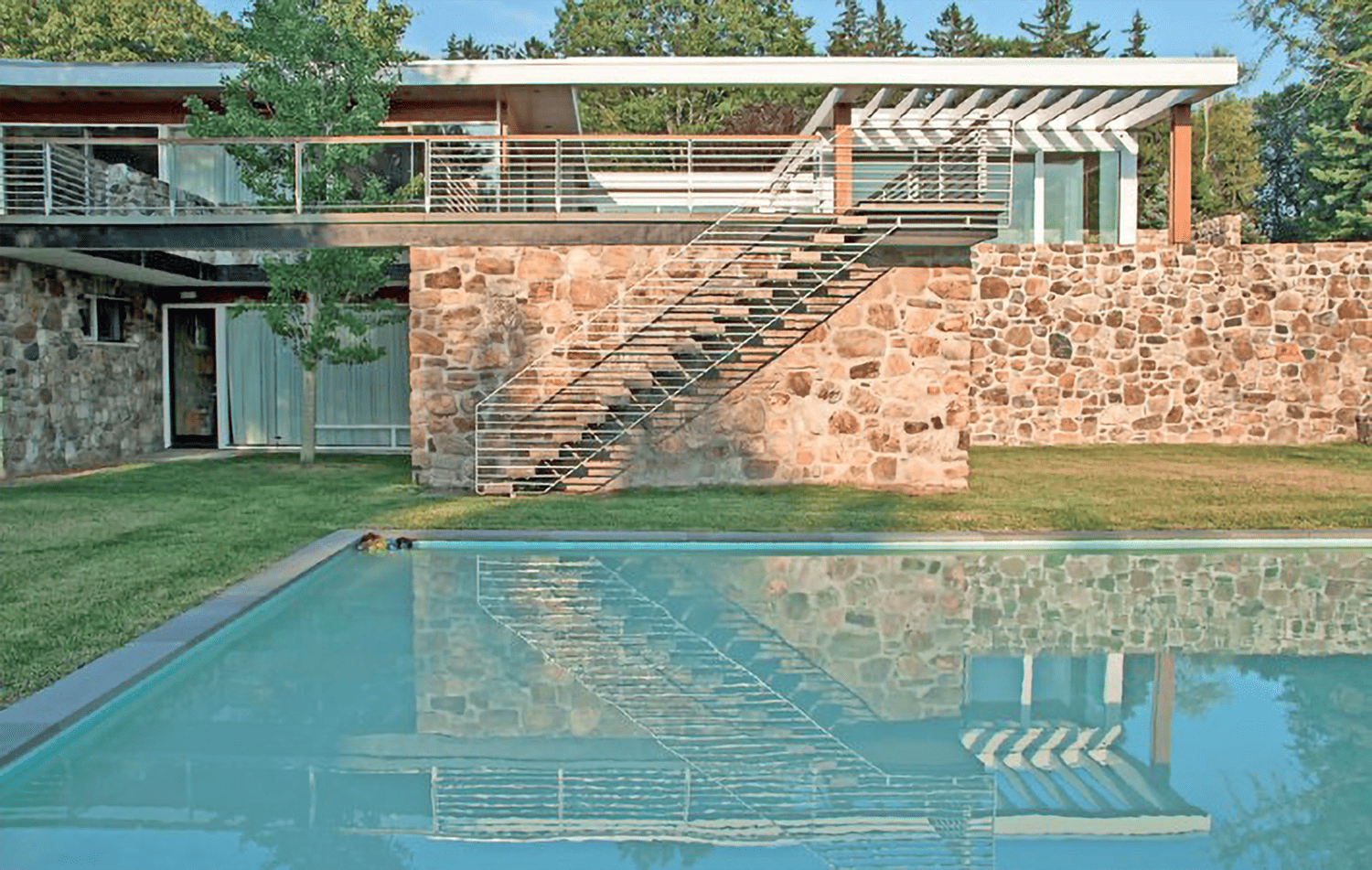 Modern house with stone wall, metal stairs, and large pool in the foreground, surrounded by trees and lawn.
