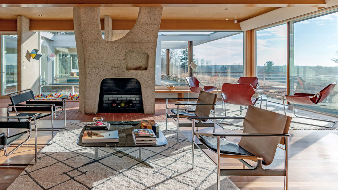 Mid-century modern living room with geometric fireplace, leather chairs, large windows, and a patterned rug.