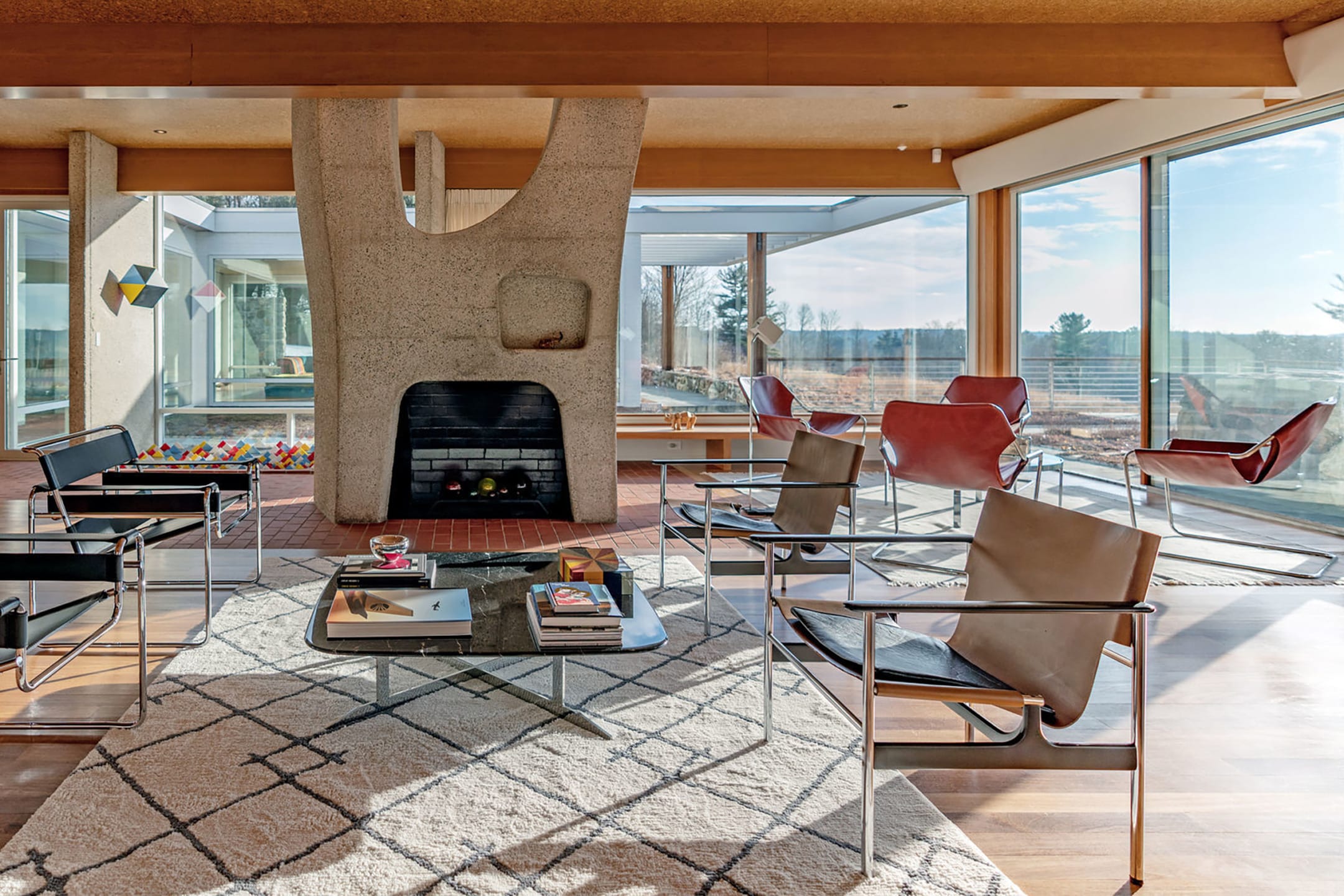 Mid-century modern living room with geometric fireplace, leather chairs, large windows, and a patterned rug.