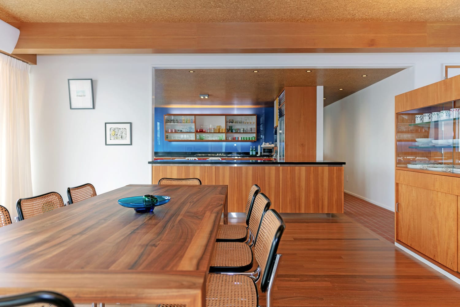 Modern kitchen and dining area with wooden table, blue accent wall, and shelves with dishes under warm lighting.