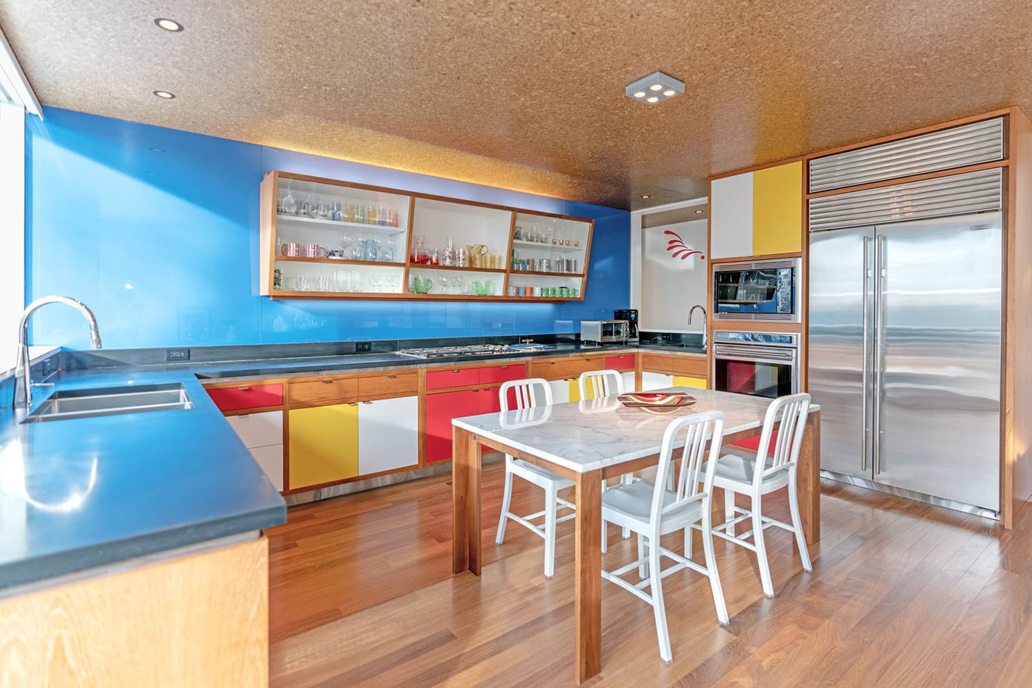 Modern kitchen with colorful cabinets, blue backsplash, and a wooden dining table with white chairs.