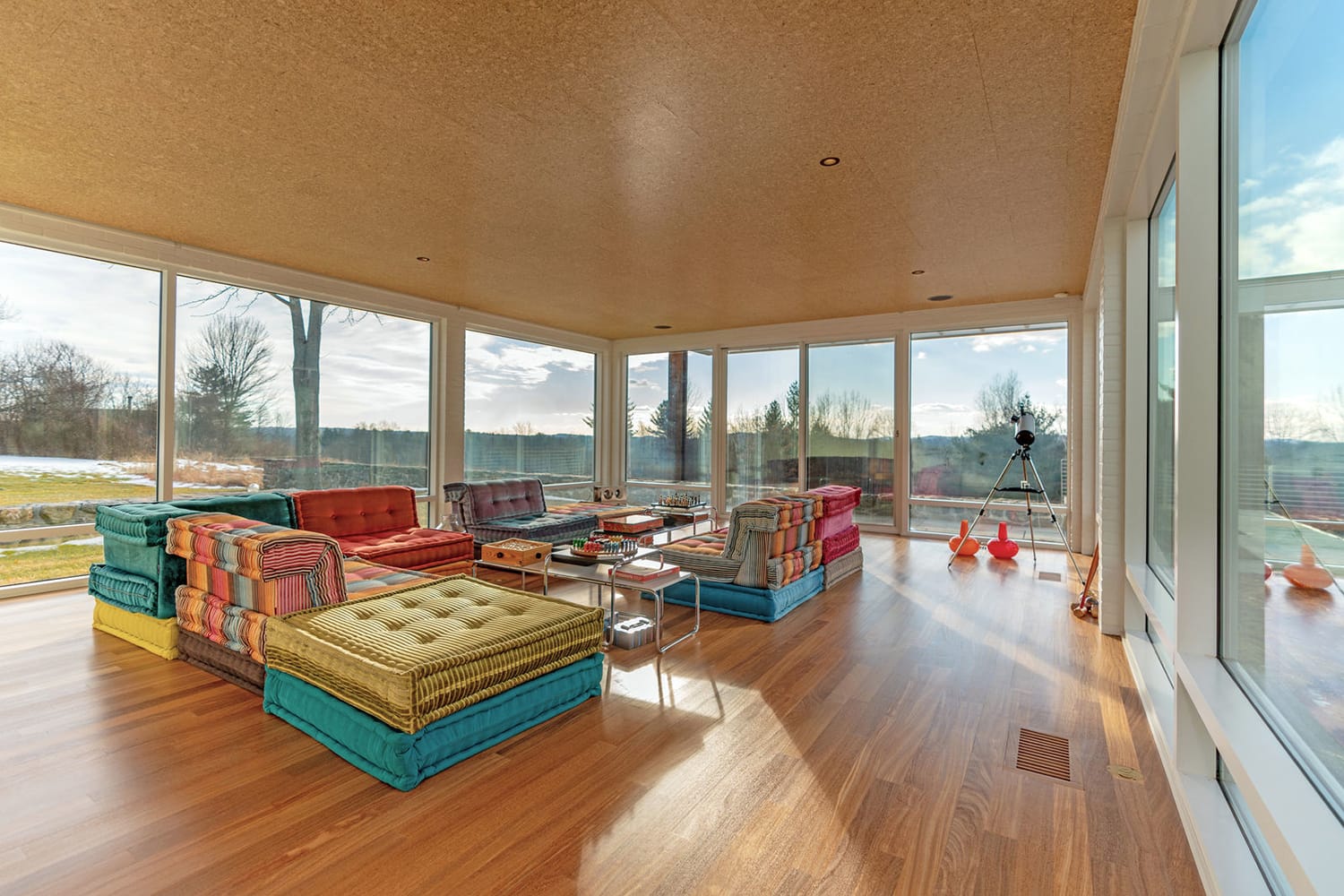Spacious sunroom with colorful modular seating, wooden floor, large windows offering a scenic outdoor view, and a telescope.
