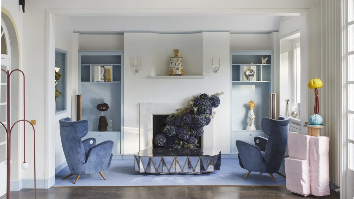 Mid-century modern living room with blue velvet chairs, floral display, geometric fireplace, and decorative shelves.