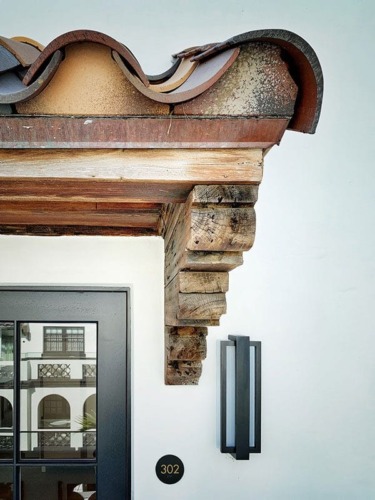 Decorative wooden corbel supporting roof overhang next to a black-framed window and modern wall sconce on a white building.