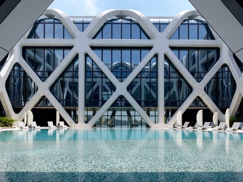 Futuristic building with geometric design, reflective glass facade, and a pool in the foreground under a clear blue sky.