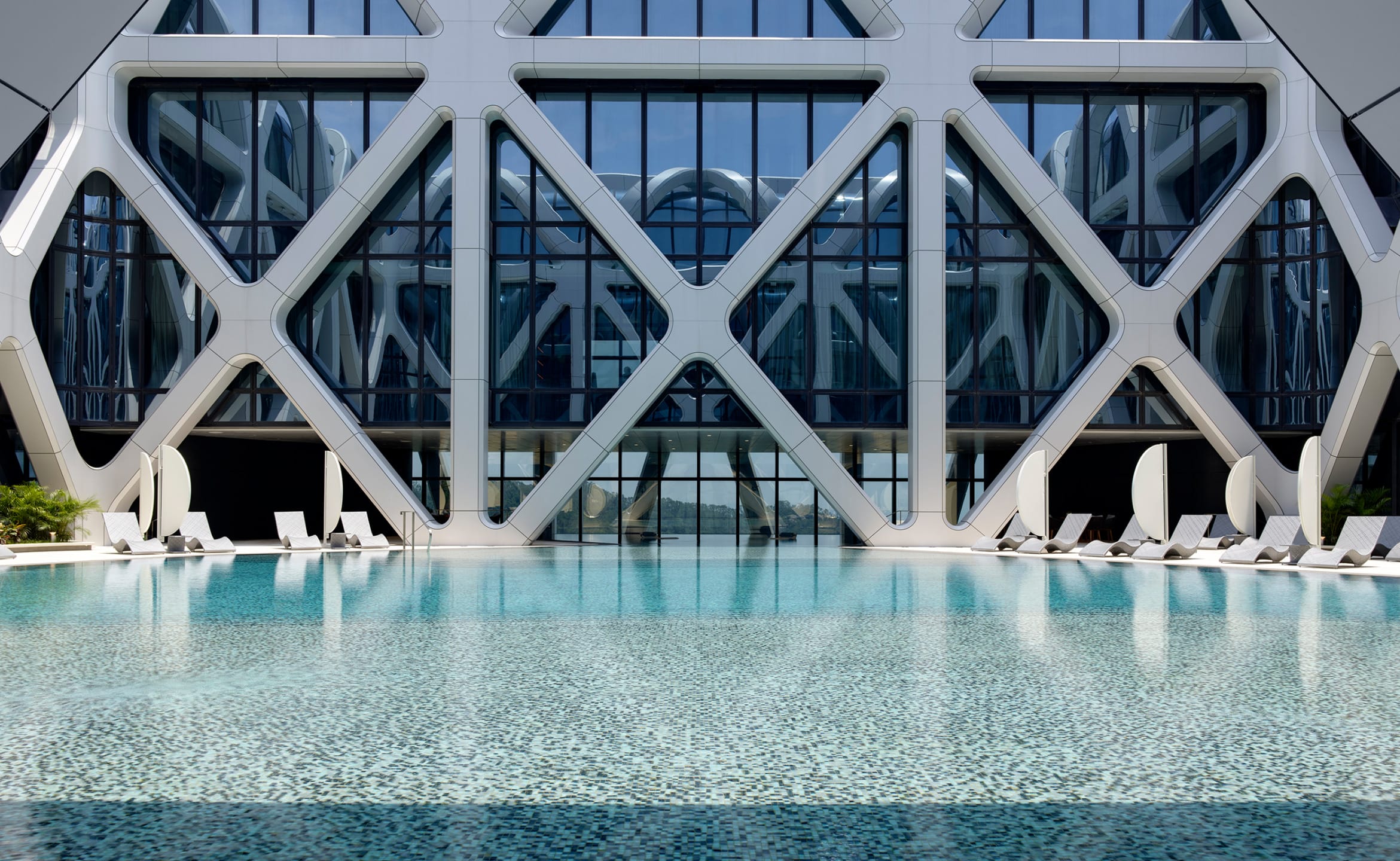 Outdoor pool with lounge chairs in front of modern building with geometric design and large glass windows.