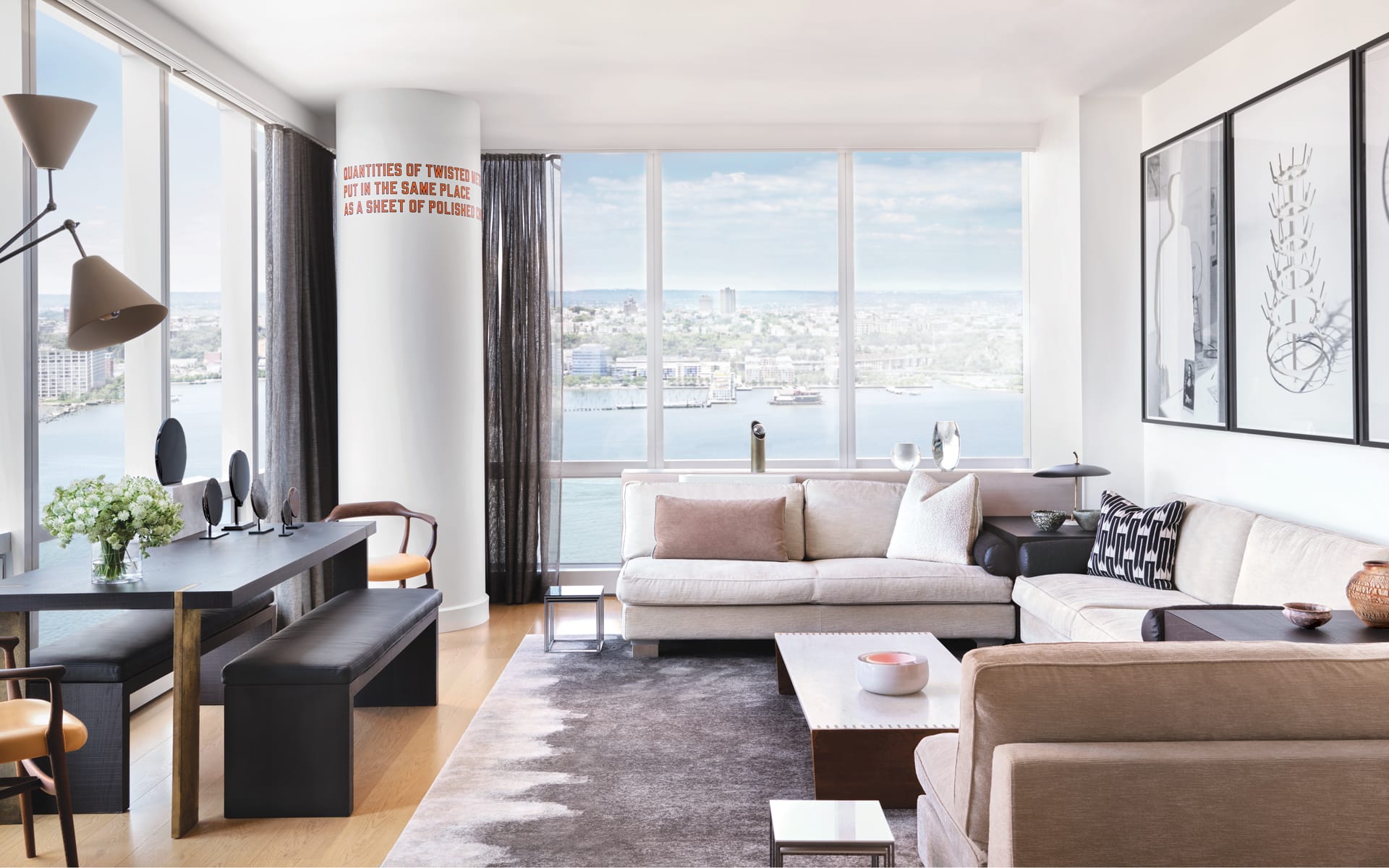 Modern living room with large windows, plush sofas, abstract art, dining area, and a view of the river and cityscape.