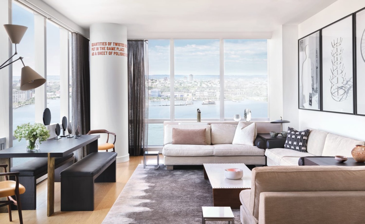 Modern living room with large windows, city and water view, stylish furniture, and contemporary art on the walls.