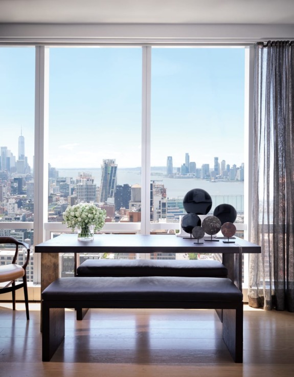 Modern dining room with city skyline view through large windows, featuring a wooden table, benches, and decorative items.