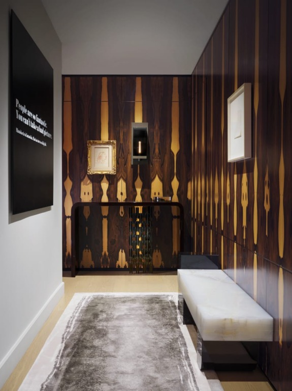 Modern interior hallway with patterned wooden wall panels, contemporary art, a white bench, and a soft rug on the floor.