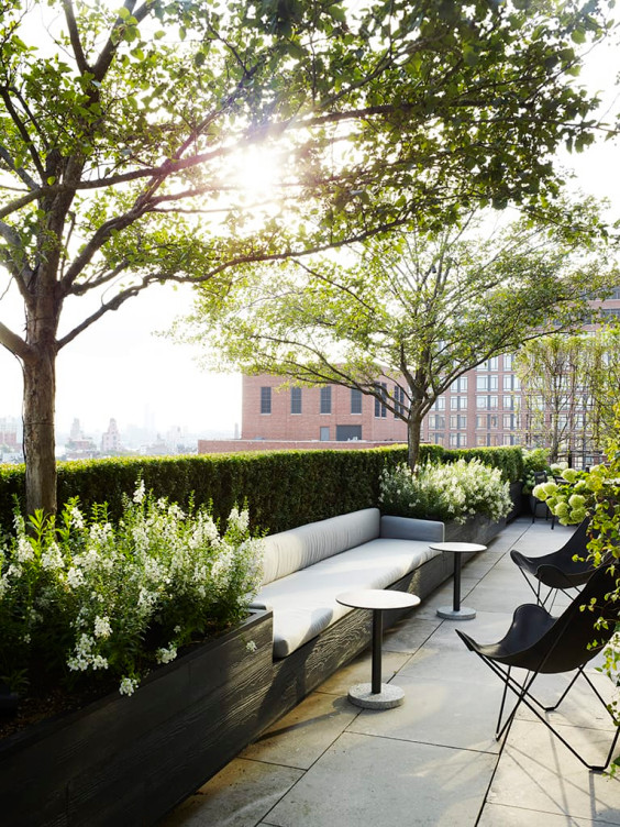 Sunny rooftop garden with seating, surrounded by trees and white flowers, creating a peaceful urban retreat.