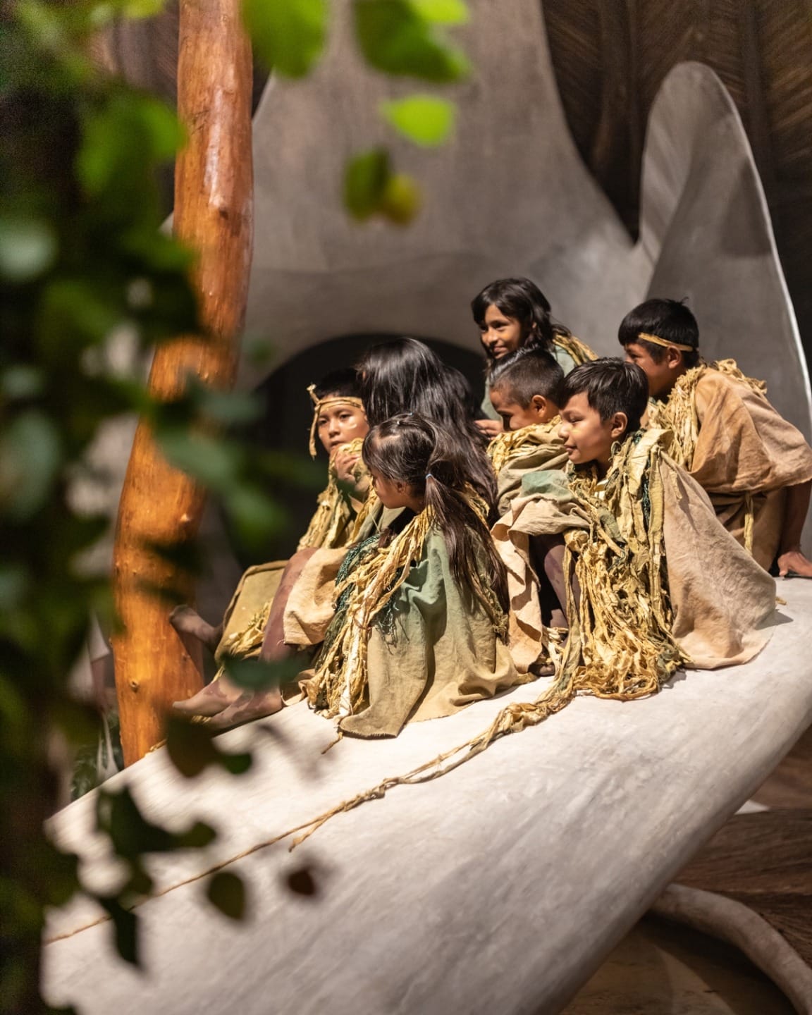 Children in traditional clothing sitting on a large object, surrounded by natural elements in an indoor setting.
