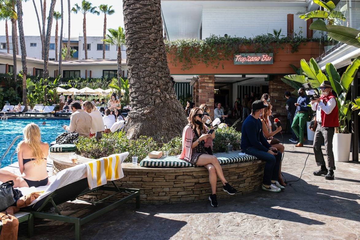 People enjoying a sunny day at a lively outdoor cafe with palm trees and a pool in the background.