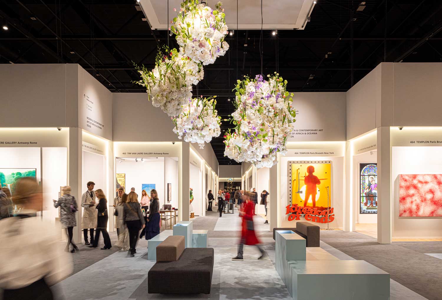 Art gallery with people viewing various colorful artworks and floral installations hanging from the ceiling.