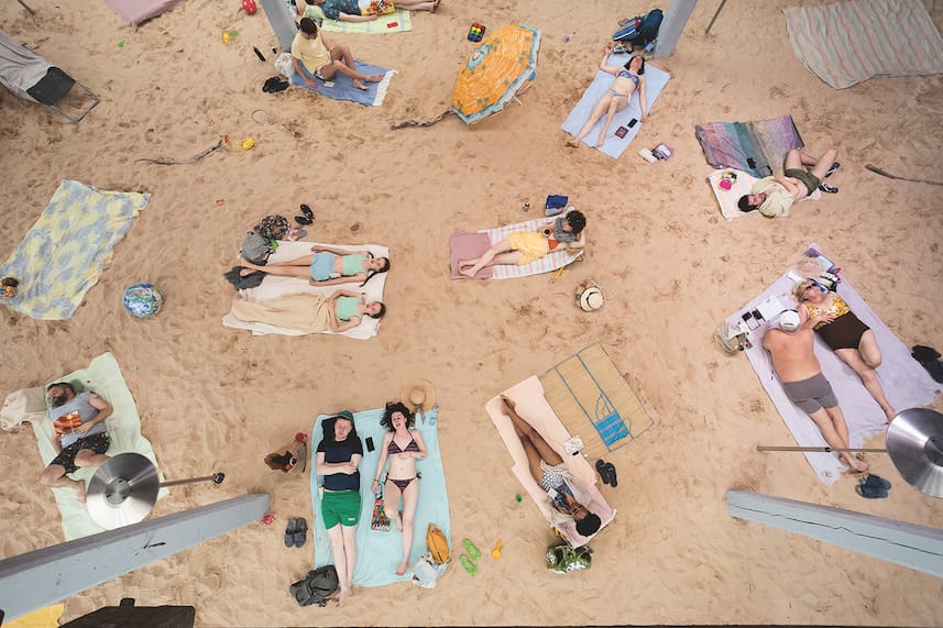 People relaxing on sandy beach with colorful towels, umbrellas, and accessories, viewed from above.