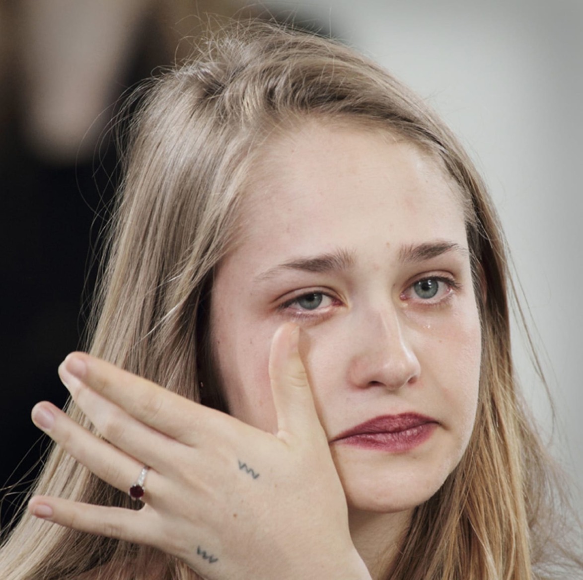 Person wiping tears from eyes while looking forward, wearing maroon lipstick, light-colored hair falling loosely around shoulders.