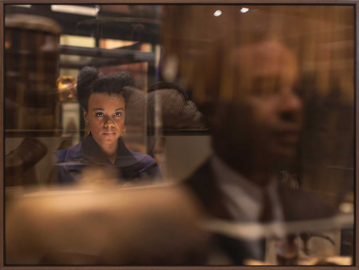Person viewed through blurred reflections, focusing intensely, surrounded by museum artifacts.