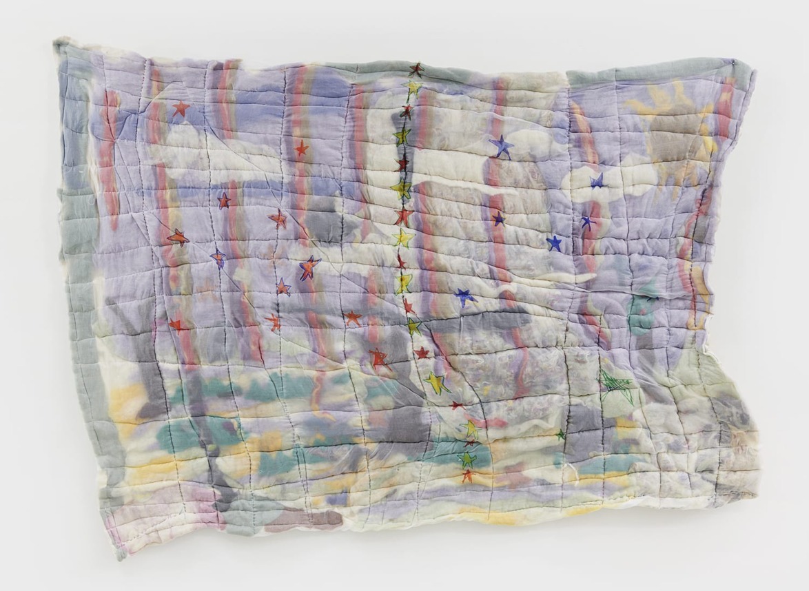 Colorful abstract quilt with stitched stars and pastel hues of purple, green, and gray hanging on a white wall.