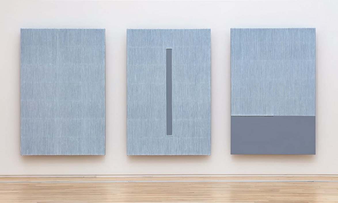 Three minimalist gray canvases with vertical lines on a gallery wall, featuring subtle texture and geometric design.