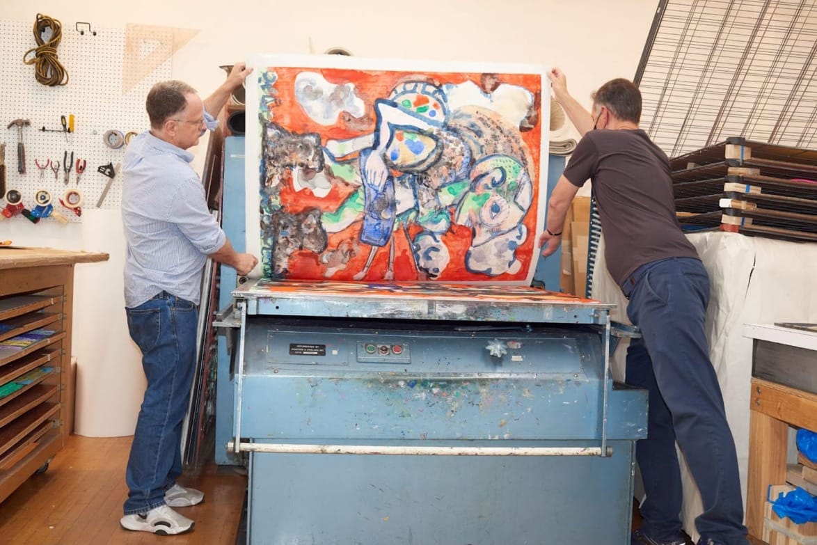Two men handling a colorful abstract painting on a large printing press in an art studio setting.