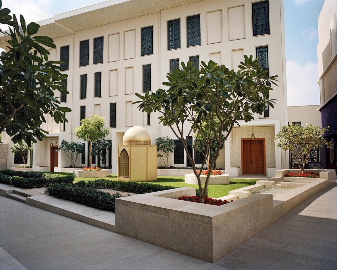 Modern courtyard with geometric architecture, green gardens, trees, and a central small dome structure.