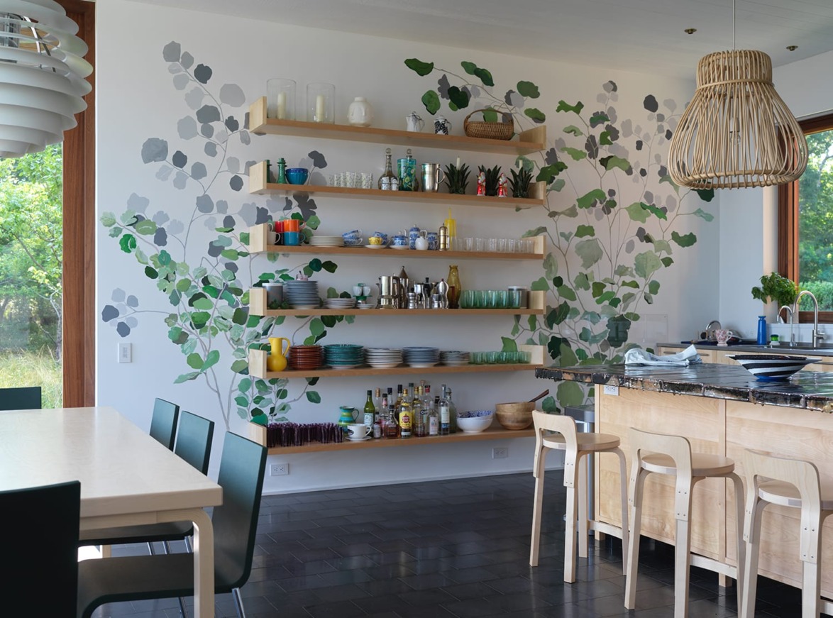 Modern kitchen with open wooden shelves, green leaf wall mural, dining table, and bar stools around an island.