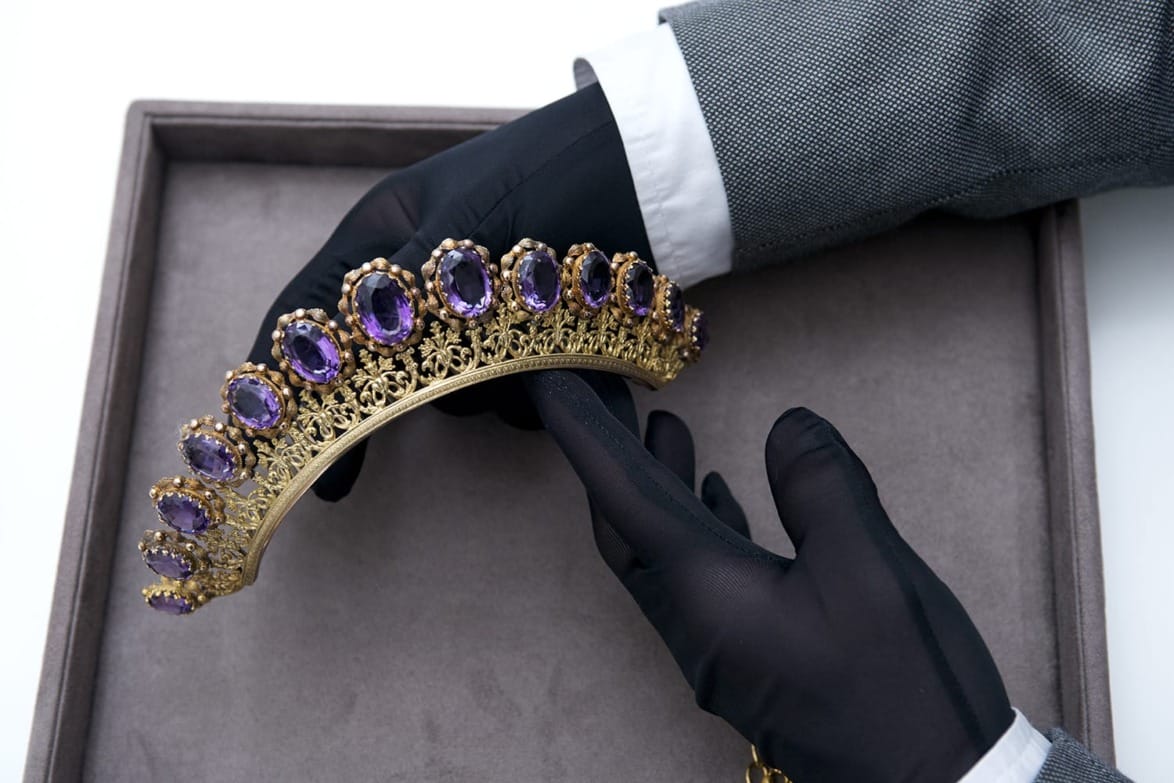 Gloved hands holding an ornate gold tiara with purple gemstones in a grey display box.