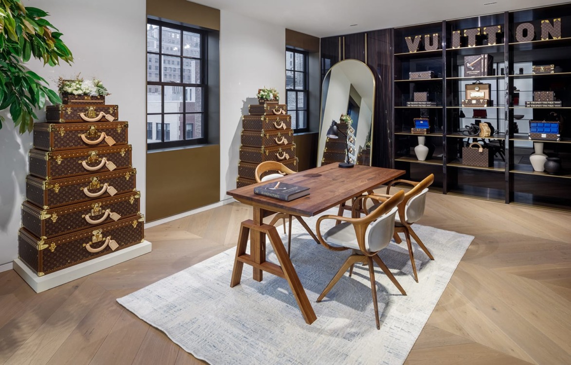 Luxury showroom with wooden table, chairs, stacked designer trunks, decorative shelving, and a large mirror.