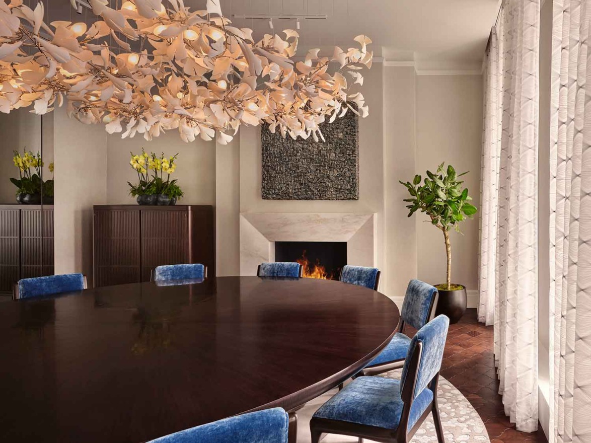 A dining room with a round wooden table, blue velvet chairs, floral chandelier, fireplace, and plants by the window.