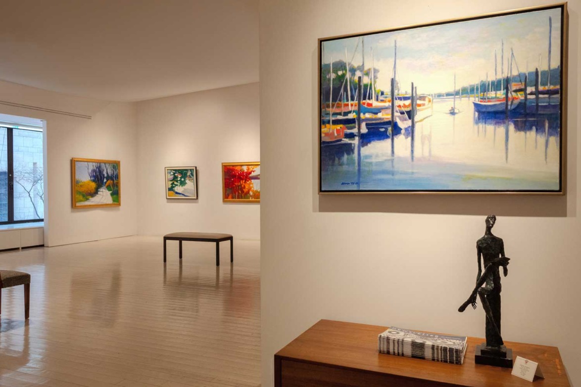 Art gallery with paintings on the walls and a sculpture on a table, featuring boats and abstract landscapes.