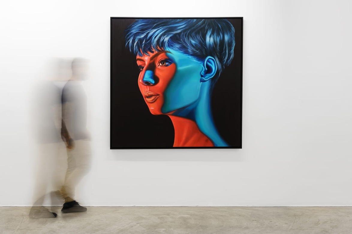 Person walking past a vibrant portrait painting with red and blue hues on a white gallery wall.