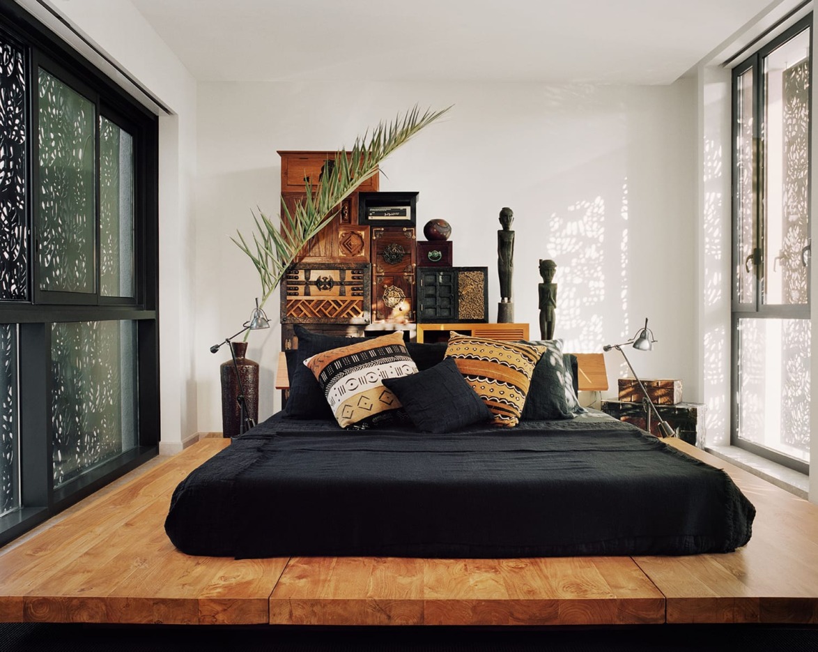 Modern bedroom with black bedding, wooden platform, decorative cushions, and ethnic decor by large windows and patterned screens.