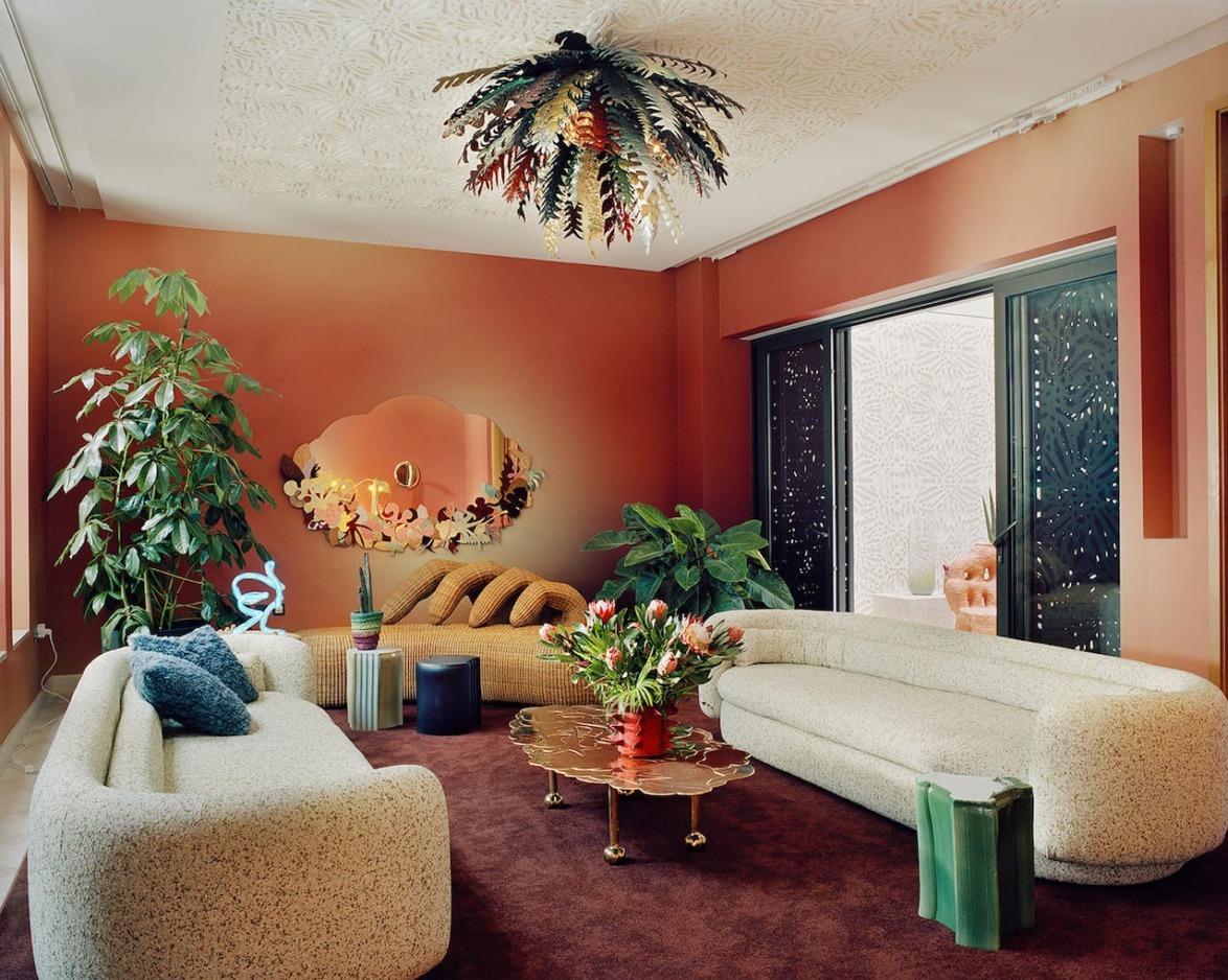 Modern living room with cozy sofas, unique leaf-shaped chandelier, large plants, and a stylish mirror on a red wall.